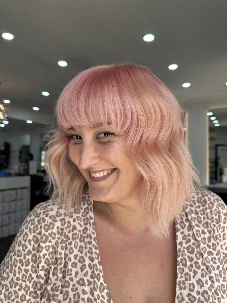 Smiling woman with shoulder-length pastel pink wavy hair and a full fringe in a hair salon.