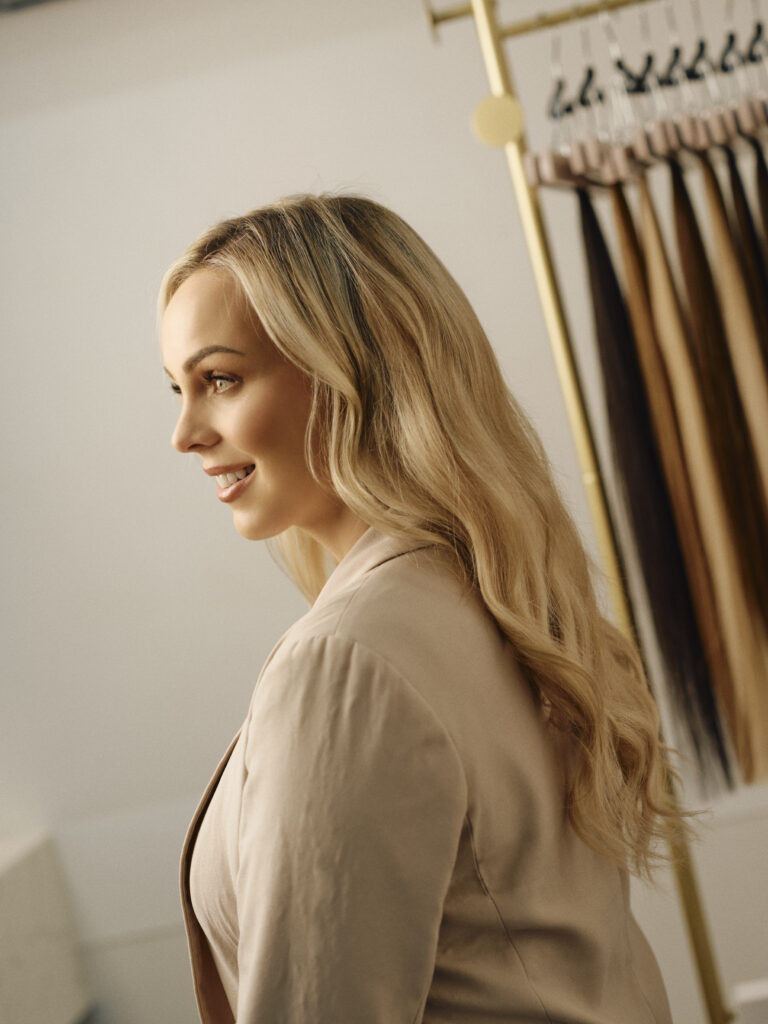Blonde client with long blended hair beside the Showpony extension colour rack at Epic Hair Designs