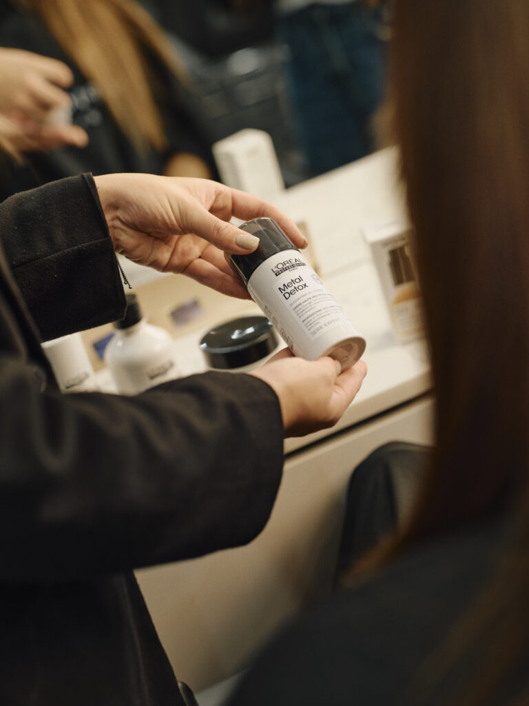 Epic Hair Designs stylist holding L'Oréal Professionnel Metal Detox High Protection Cream during a client consultation.