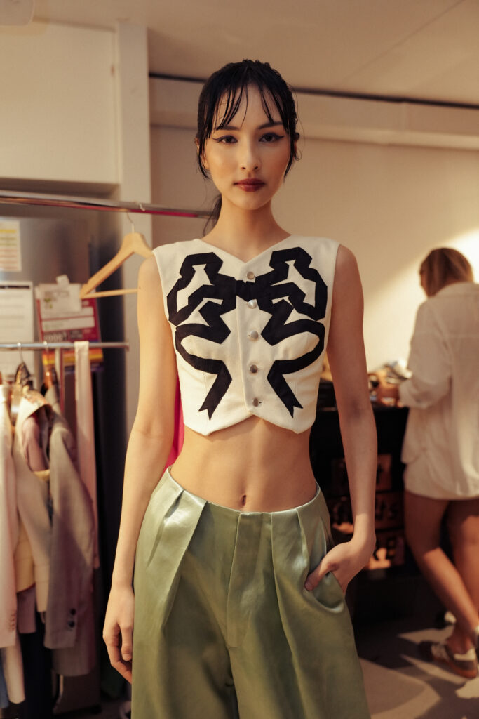 Model backstage at Brisbane Fashion Month wearing a graphic bow crop top and sage trousers with a sleek wet-look updo.