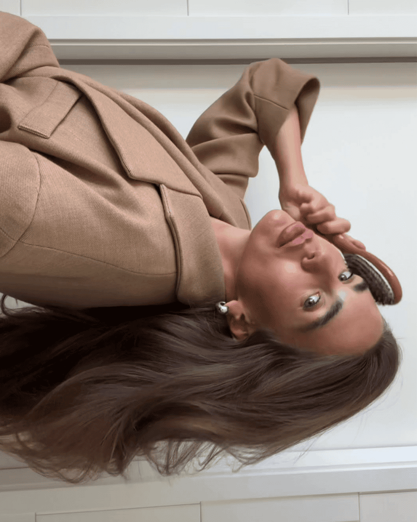 Woman brushing her long, glossy brunette hair while wearing a camel-coloured blazer.