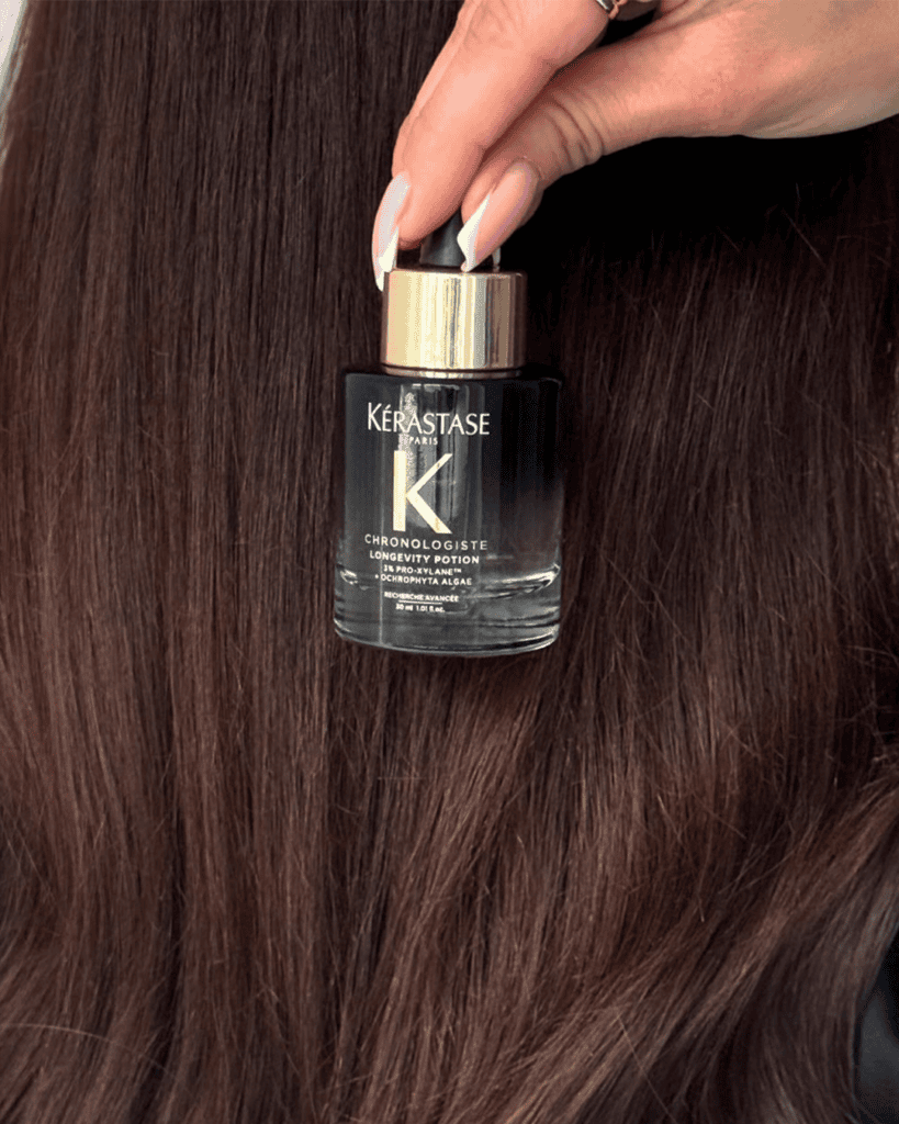 Hand holding a bottle of Kérastase Chronologiste Longevity Potion against long, glossy brunette hair.